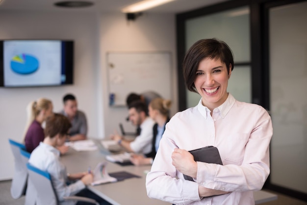 retrato de una joven mujer de negocios hispana con una tableta en el moderno interior de la oficina de negocios de inicio, grupo de personas en una reunión de equipo borrosa en el fondo