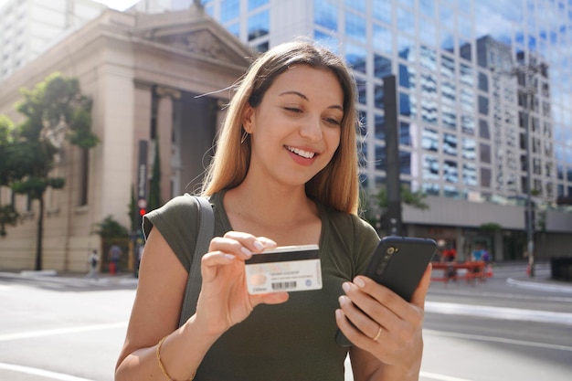 Retrato de una joven mujer de negocios mirando su teléfono con tarjeta de crédito en la avenida del distrito financiero