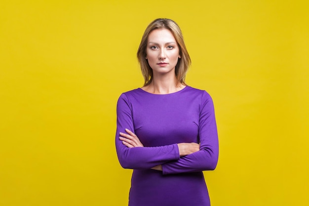 Retrato de una joven mujer de negocios segura de sí misma con un elegante vestido morado ajustado de pie con las manos cruzadas con un aspecto exitoso, inteligente y profesional en un estudio interior aislado en un fondo amarillo