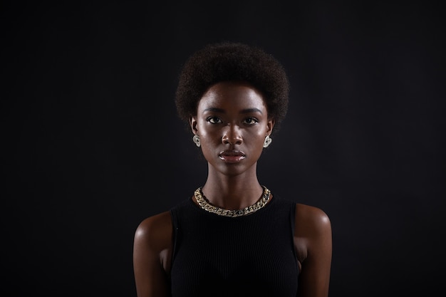 Retrato de joven mujer negra elegante seria. Hermosa chica afroamericana de pelo oscuro rizado usa vestido negro con joyas de oro y mirando a cámara. Sesión de estudio aislado sobre fondo oscuro.