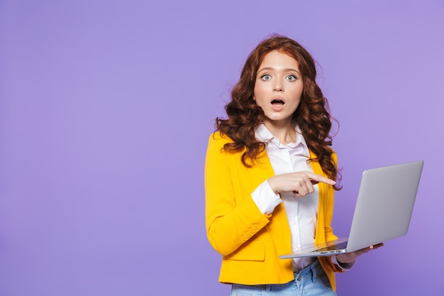Retrato de una joven mujer pelirroja bastante sorprendida de pie sobre violeta, usando la computadora portátil