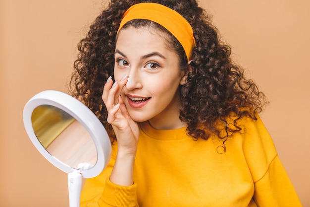 Retrato de joven mujer rizada con crema para la piel en un espejo aislado sobre fondo beige. Concepto de cuidado de la piel.