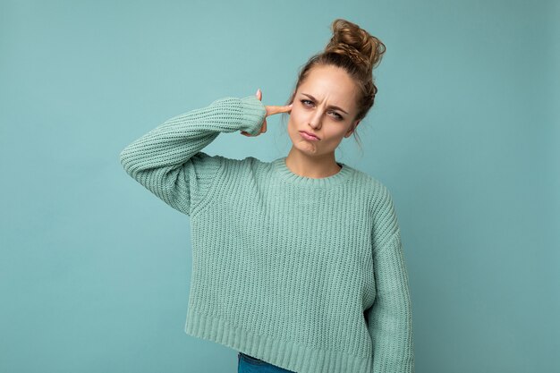 Retrato de joven mujer rubia atractiva emocional triste triste con emociones sinceras vistiendo jersey azul casual aislado sobre fondo azul con espacio vacío y haciendo pistola de dedo cerca de la cabeza.