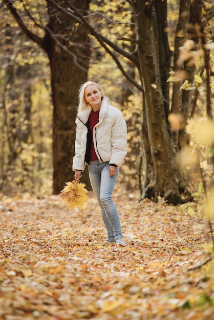 Retrato de una joven mujer rubia en el bosque de otoño, con un ramo de hojas amarillas en sus manos.