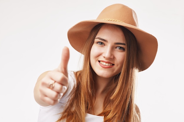 Retrato de una joven mujer rubia sonriente con sombrero beige mostrando el pulgar hacia arriba