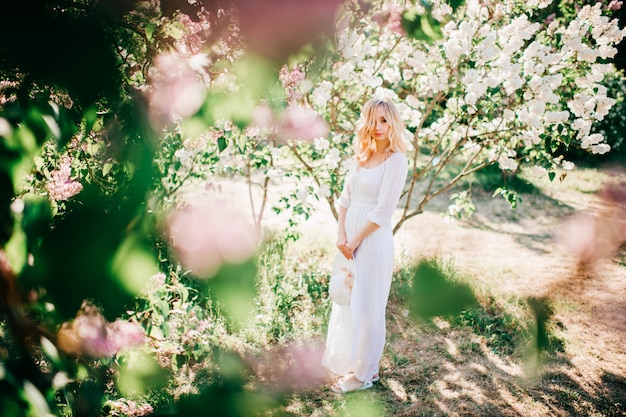 Retrato de joven mujer rubia en vestido blanco elegante en verano floreciente jardín lila.