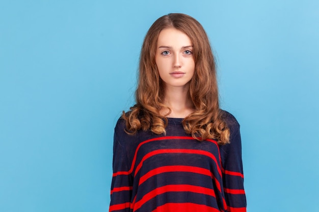 Retrato de una joven mujer seria de cabello ondulado que usa un suéter de estilo casual a rayas de pie con una estricta expresión mandona, sin sonreír mirando la cámara. Disparo de estudio interior aislado sobre fondo azul.