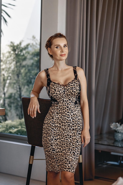 Retrato de una joven mujer sexy y elegante que lleva un vestido ajustado con estampado de leopardo y posa junto a una silla en su casa. Concepto de vida rica