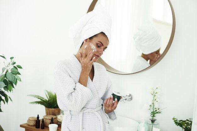 Retrato de una joven mujer sonriente aplicando crema hidratante en el rostro en el baño. Cosmetología. Belleza y Spa.