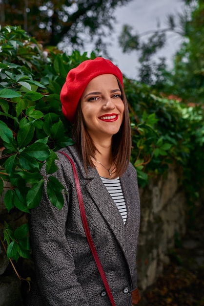 Retrato de una joven mujer sonriente en boina roja apoyado en una valla de piedra cubierta de vegetación