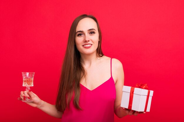Retrato de una joven mujer sonriente con un elegante vestido rosa sosteniendo una copa de champán y una caja de regalo Fiesta de Navidad