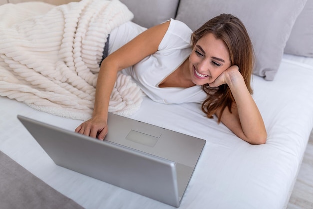 Retrato de una joven mujer sonriente sentada con un portátil en la cama. Feliz hermosa mujer casual trabajando en un portátil tendido en la cama de la casa. Mujer freelance trabajando en su computadora portátil en casa