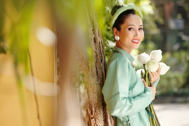 Retrato de joven mujer vietnamita feliz en vestido ao dai y headser tradicional sosteniendo flores de loto cuando está de pie en el parque de la ciudad