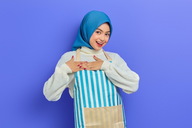 Retrato de una joven musulmana asiática sonriente con hiyab y delantal rayado de pie con las manos dobladas en el corazón y mirando la cámara aislada en un fondo morado