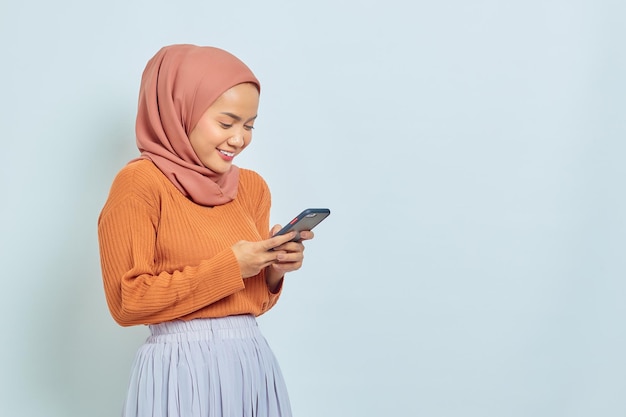 Retrato de una joven musulmana asiática sonriente con suéter marrón y hiyab usando un teléfono móvil aislado en un concepto de estilo de vida musulmán de fondo blanco