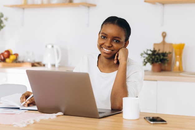 Retrato de una joven negra sentada en la cocina y trabajando en una laptop desde casa