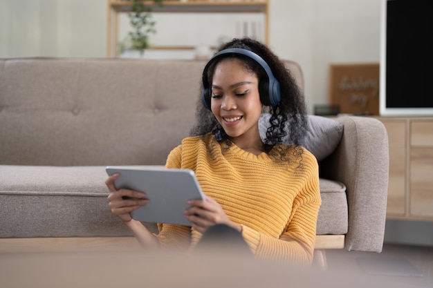 Retrato de una joven negra sonriente con auriculares inalámbricos sosteniendo una tableta digital sentada en la sala de estar