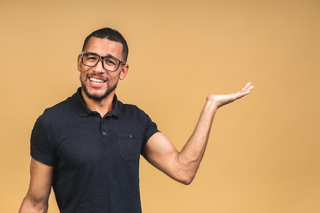 Retrato de un joven negro afroamericano feliz con una sonrisa casual señalando a un lado con el dedo mirando a la cámara con expresión de cara emocionada aislada sobre fondo beige