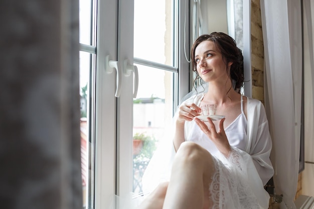 El retrato de una joven novia hermosa en un bata se sienta en una ventana y espera al novio