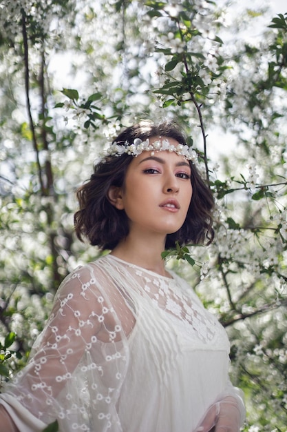 Retrato de una joven novia hermosa vestida de blanco parada al lado de un cerezo floreciente en primavera una corona en la cabeza