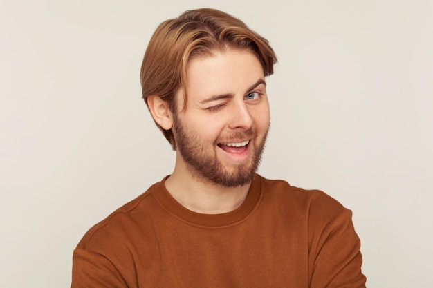 Retrato de un joven optimista y feliz con el pelo y la barba pulcros con una sudadera guiñando un ojo a la cámara