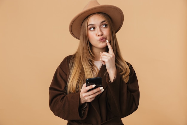 Retrato de joven optimista con sombrero pensando y sosteniendo teléfono celular aislado en beige
