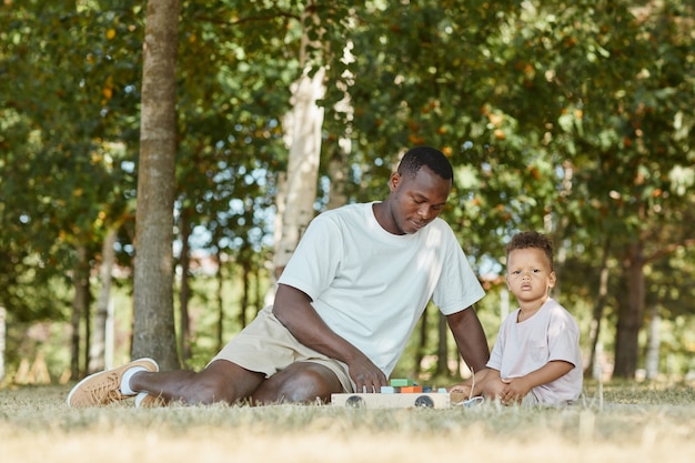 Retrato de joven padre afroamericano jugando con su hijo lindo en el parque mientras está sentado en la hierba copia s ...