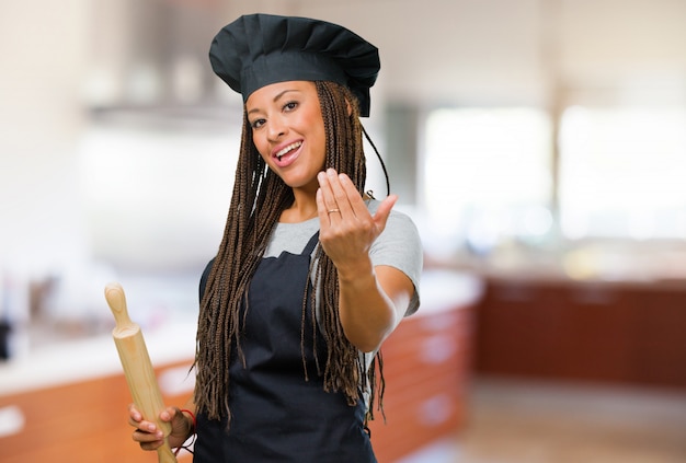 Retrato de una joven panadera negra que invita a venir, confiada y sonriente haciendo un gesto con la mano, siendo positiva y amigable.