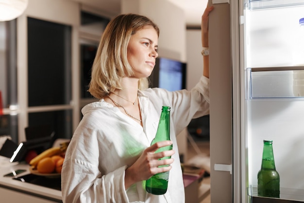 Retrato de una joven parada en la cocina por la noche y mirando cuidadosamente en la nevera abierta mientras sostenía cerveza en la mano en casa