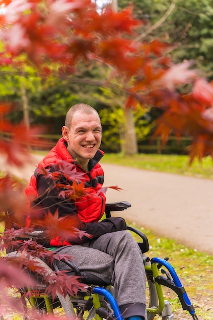 Retrato de un joven paralizado con un chaleco rojo en un parque público de la ciudad Sentado en la silla de ruedas junto a hojas rojas