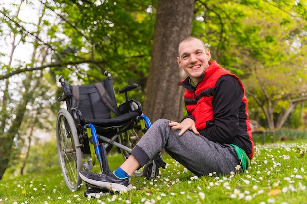 Retrato de un joven paralizado sentado en el césped junto a la silla de ruedas sonriendo en la primavera en un parque natural