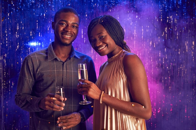 Retrato de joven pareja afroamericana sosteniendo copa de champán y sonriendo a la cámara mientras disfruta de la fiesta en la discoteca, espacio de copia