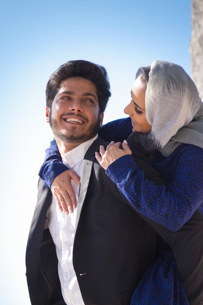 Retrato de joven pareja árabe amorosa abrazándose. Hermosa mujer con pañuelo en la cabeza abrazando el cuello del hombre feliz desde la espalda y tiernamente mirando el uno al otro. Concepto de felicidad, relación y romance.