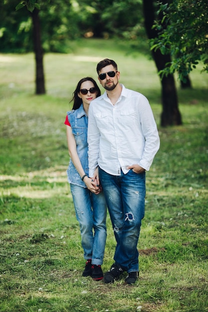 Retrato de una joven pareja feliz enamorada sonriendo y abrazándose en el jardín Dulces amantes con elegantes gafas de sol oscuras y ropa informal posando y mirando la cámara durante el día en el parque de verano