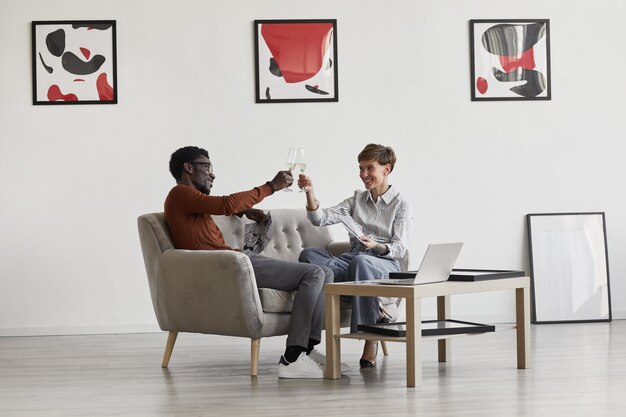 Retrato de joven pareja de raza mixta disfrutando de champán mientras está sentado en el sofá en la habitación decorada con arte moderno,