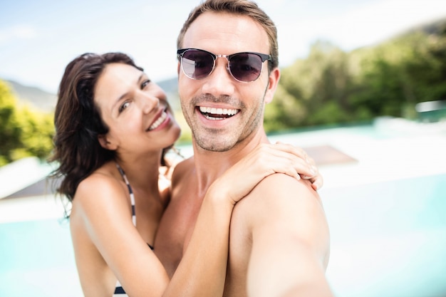 Retrato de joven pareja sonriendo junto a la piscina en un día soleado