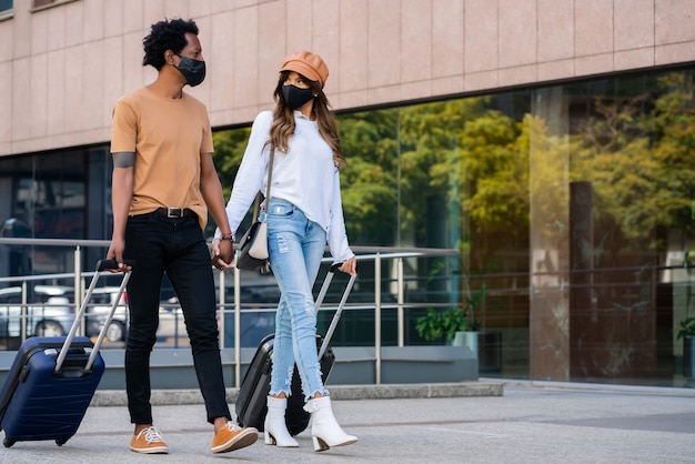 Retrato de joven pareja de turistas con máscara protectora y maleta mientras camina al aire libre en la calle. Concepto de turismo. Nuevo concepto de estilo de vida normal.