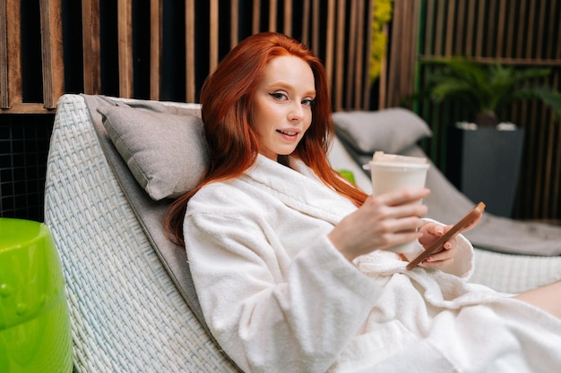 Retrato de una joven pelirroja con albornoz relajándose con un smartphone tumbado en una tumbona junto a la piscina y bebiendo un cóctel después de un tratamiento de belleza en el salón de spa