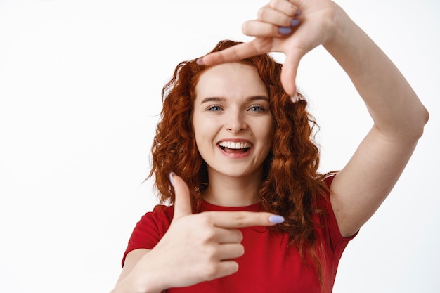 Retrato de joven pelirroja creativa mostrando gesto de marco de mano, tomando una foto de usted con cara sonriente feliz, de pie en camiseta contra la pared blanca