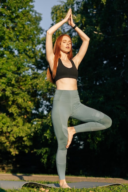 Retrato de una joven pelirroja meditativa haciendo yoga en pose de árbol levantando las manos en pose de Namaste