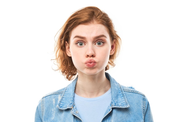 Retrato de una joven pelirroja con las mejillas hinchadas y la cara graciosa aislada sobre un fondo blanco de estudio Concepto de humor Boca inflada con aire Expresión loca