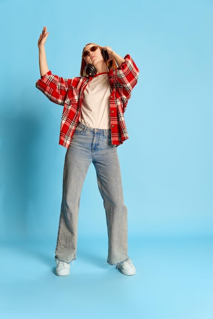 Retrato de una joven pelirroja con un pantalones casual a cuadros escuchando música con auriculares y bailando sobre fondo azul.