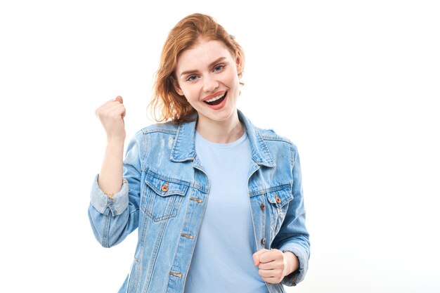 Retrato de una joven pelirroja sobre un fondo blanco aislado en jeans. Mira a la cámara tomados de la mano frente a ti en un puño