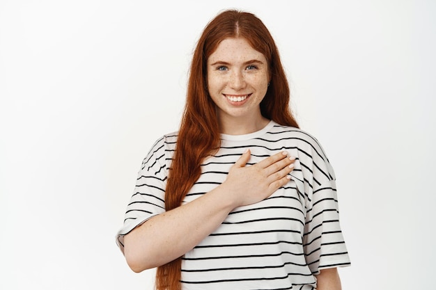 Retrato de una joven pelirroja sonriente sosteniendo la mano en el corazón y mirando a la cámara agradecida, agradeciéndole, jurando, comprometiéndose con algo, mostrando devoción, fondo blanco