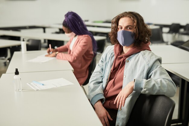 Foto retrato de joven de pelo largo con máscara en la escuela y mirando a la cámara mientras está sentado en el escritorio en el aula, espacio de copia