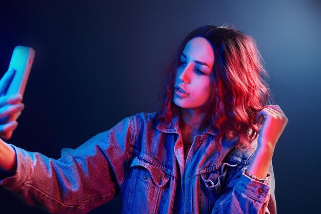 Retrato de una joven con el pelo rizado que se hace selfie usando un teléfono en neón rojo y azul en el estudio