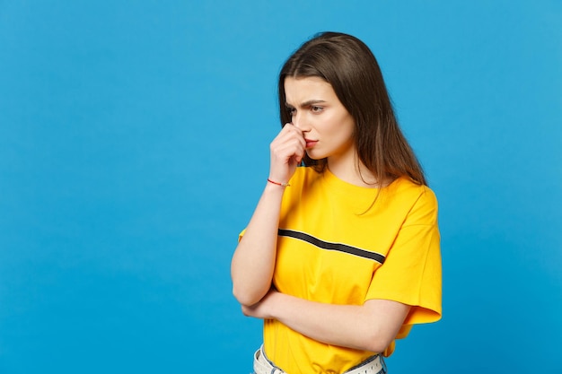 Retrato de una joven pensativa disgustada con ropa informal vívida mirando a un lado manteniendo la mano cerca de la cara aislada en un fondo de pared azul brillante en el estudio. Concepto de estilo de vida de las personas. Simulacros de espacio de copia.