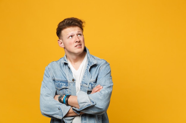 Retrato de un joven perturbado con ropa informal de denim mirando hacia un lado, con las manos cruzadas aisladas en un fondo amarillo anaranjado. Gente emociones sinceras concepto de estilo de vida. Simulacros de espacio de copia.