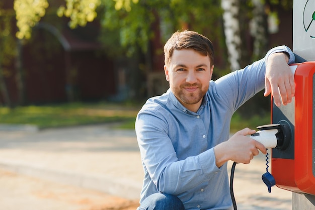 Retrato de un joven de pie con un cable de carga cerca de la estación de carga Concepto de cargadores de automóviles rápidos para el hogar