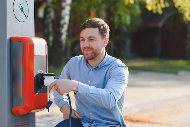 Retrato de un joven de pie con un cable de carga cerca de la estación de carga Concepto de cargadores de automóviles rápidos para el hogar